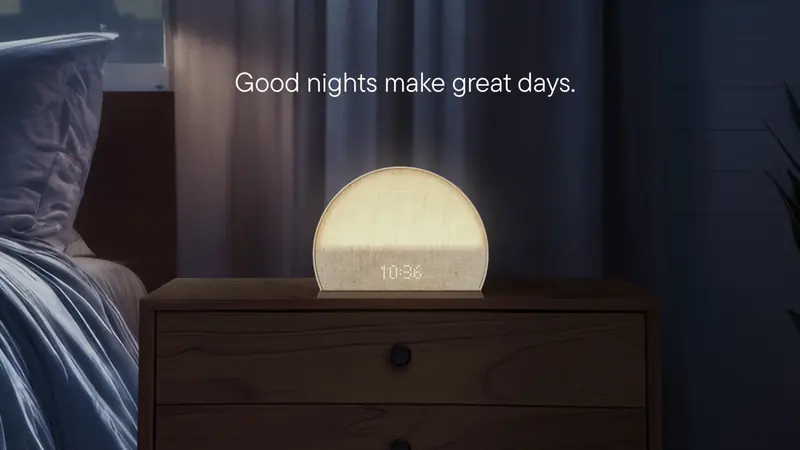Le même réveil diffusant une lumière douce sur une table de chevet, dans une chambre plongée dans la pénombre, avec l’accroche en anglais « Good nights make great days » (les bonnes nuits font les belles journées).
